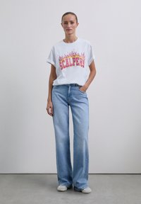 Donna in piedi con una mano in tasca, che indossa una maglietta bianca con stampa di fiamme "SCALPERS", jeans a gamba larga di colore azzurro chiaro e sneakers bianche.