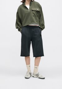 Personne porte un pull court vert olive, un short noir jusqu’aux genoux, des chaussettes Nike beiges et des baskets blanches avec « 90 » inscrit dessus.