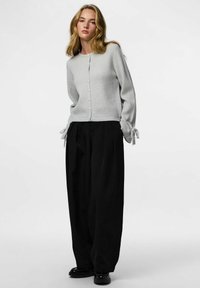 Heled hall ribane kardigan, millel on ees nupud ja seotud varrukad. Sobitatud lühikeste mustade pükstega ja mustade kingadega.