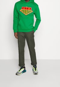 Sudadera de felpa verde con el logotipo bordado de "AIR JORDAN", combinada con pantalones chinos oliva y zapatillas multicolores con acentos neón.