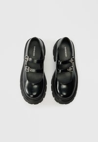 Sapatos Mary Jane em couro preto, com dois atacadores ajustáveis com fivelas prateadas, bico redondo e solas de borracha grossa.