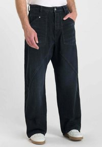 Donkerblauwe denim wijde broek met hoge taille, voorzien van frontzakken en unieke diagonale stiksels. Gecombineerd met witte sneakers.