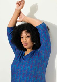 Sininen paita, jossa on eloisa kuvio violetteina sävyinä, rento leikkaus, kolmivartiset hihat ja syvä V-aukko.