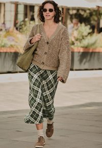 Brauner, strukturierter Cardigan mit großen Knöpfen, grüne karierte Schlaghosen, braune Sneaker und eine olivgrüne Tasche; Sonnenbrille vervollständigt den Look.