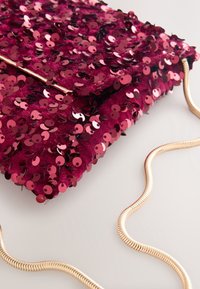 Petit sac rectangulaire bordeaux couvert de sequins brillants et attaché à une fine chaîne dorée reposant sur une surface claire.