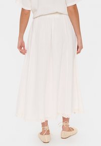 Jupe maxi blanche avec un design fluide, un tissu texturé et une bordure en dentelle festonnée au bas. Portée avec des chaussures espadrilles en beige à lanières.