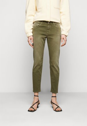 Persona con pantalones ajustados verde oliva, chaqueta color crema y tacones negros con tiras y dedos descubiertos, de pie sobre un fondo liso.