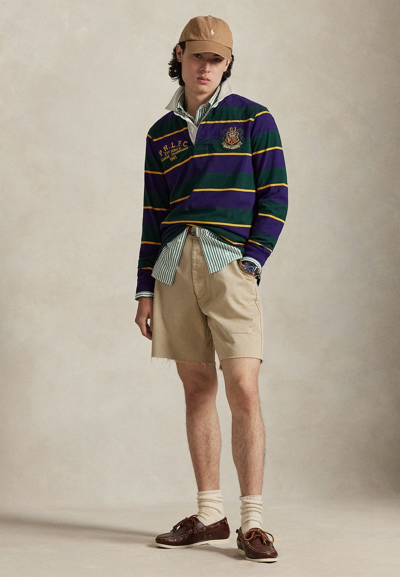Chemise de rugby multicolore avec des rayures violettes et vertes, short beige, mocassins marron et casquette beige. Superposée avec une chemise rayée claire.