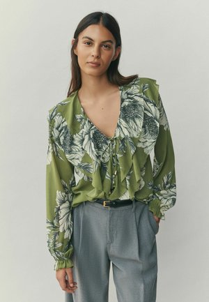 Vihreä kukkakuvioinen pusero, jossa liehutitettu malli, syvä pääntie ja pitkät hihat. Yhdistetty harmaisiin housuihin, joissa on hienovarainen tekstuuri ja vyö.