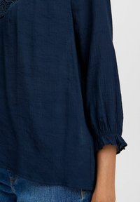 Navy blue blouse with a textured fabric, featuring long sleeves with ruffled cuffs and a loose fit. Simple design, no visible patterns or hardware.