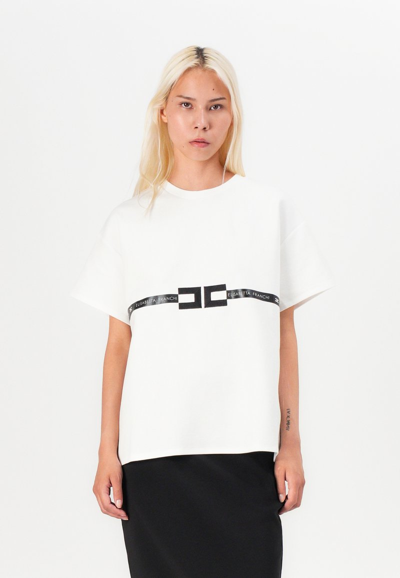 White short-sleeve t-shirt made of soft fabric, featuring a black logo band across the chest, paired with a black skirt.