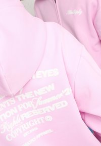Sweatshirt rose avec des détails en texte blanc, présentant une texture lisse et un design décontracté. Le texte comprend des informations sur le copyright et le branding.