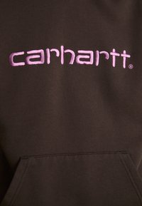 Bruine capuchontrui met een voorzak. Kenmerkt zich door een geborduurd "carhartt" logo in lichtroze. Het materiaal lijkt zacht met een losse pasvorm.