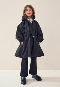 Trench negru cu glugă, cu manșete elastice și talie ajustabilă, asortat cu pantaloni negri evazați și pantofi negri. Textură netedă a țesăturii.