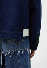 Tummansininen farkkutakki, jossa on valkoinen etiketti ja vihreä korostus; yhdistetty kuluneisiin sinisiin farkkuihin, joissa on risaiset reunat.