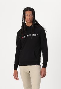 Zwarte hoodie met het "DENIM PROJECT" logo in het wit, voorzien van een voorzak, een verstelbare capuchon en geribbelde manchetten. Draagt met lichtgekleurde broek.