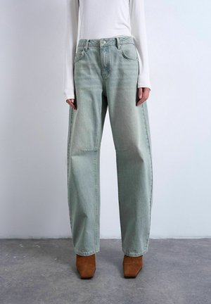 Persoon draagt losse lichtgroene spijkerbroek, wit shirt met lange mouwen en bruine laarzen met vierkante neus, staand op een grijze vloer.