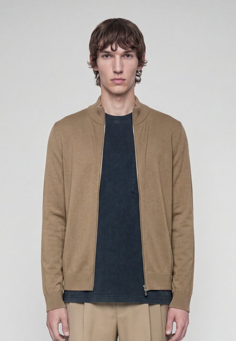 Jeune homme portant un pull zippé beige par-dessus une chemise noire et un pantalon beige, debout devant un fond uni gris clair.