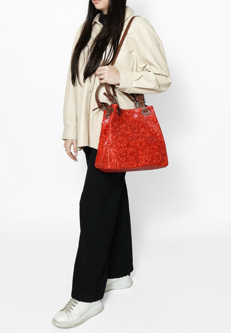 Sac à main en cuir rouge avec motif floral en relief, doté de poignées en cuir brun et de quincaillerie en argent. Modèle associé à une chemise crème et un pantalon noir.