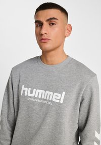 Młody mężczyzna z krótkimi włosami, ubrany w szary sweter z napisem "hummel SPORTSWEAR SINCE 1923" na piersi, na jednolitym tle.