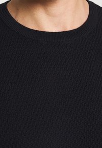 Musta neulepaita, jossa on teksturoitu kangas, resorikaulus ja vartalonmyötäinen istuvuus, esittelee pehmeän, kuvioidun pinnan.