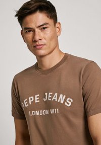 Rudas medvilninis marškinėliai su užrašu "PEPE JEANS" ir "LONDON W11" balta spalva, su apvaliu kaklu, trumpomis rankovėmis ir lygiu paviršiumi.