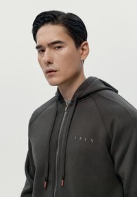 Tmavošedá mikina na zips s jemnou textúrou, so štítkom na hrudi, sťahovacími šnúrkami a kontrastnými červenými akcentmi na koncoch.