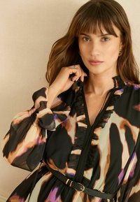 Vzdušné čierne šaty s abstraktnými hnedými, fialovými a béžovými vzormi, s volánovým golierom, naškrobenými rukávmi a čiernym opaskom.