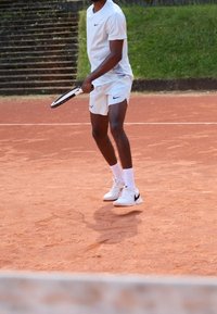 Weißes Sportoberteil und Shorts mit schwarzen Akzenten, hält einen Tennisschläger auf rotem Sandplatz. Weiße und schwarze Turnschuhe, fokussierte Haltung.