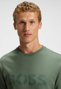 Grøn sweatshirt lavet af en blanding af bomuld. Har ribbet rund hals og præget "BOSS" logo på forsiden med et struktureret mønster.