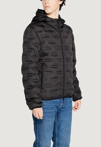 Giubbotto piumino nero con superficie testurizzata, zip completa e cappuccio. Presenta tasche laterali con zip e polsini elasticizzati. Indossato sopra un paio di jeans blu.