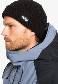 Berretto in maglia nera con superficie testurizzata e logo applicato, indossato con un giubbotto bicolore grigio e nero dotato di colletto alto e cordini.