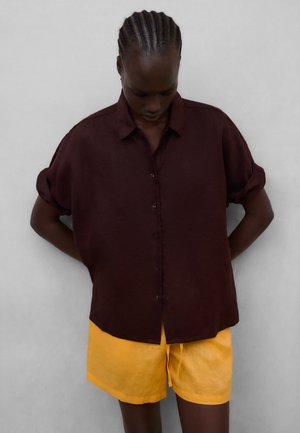 Person med fletninger iført en mørk bordeaux skjorte med knapper og optrukne ærmer samt lyse gule shorts foran en grå baggrund.