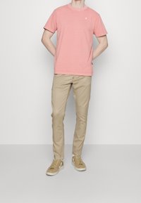 Jasnoróżowa koszulka z krótkim rękawem, beżowe spodnie o dopasowanym kroju oraz beżowe trampki z białymi dodatkami, stojące na jednolitym tle.