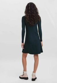 Femme avec des cheveux bouclés portant une robe noire à manches longues et des bottes de combat beiges, se tenant de dos contre un fond uni.