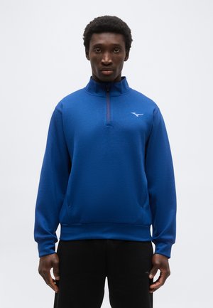 Άντρας που στέκεται με πρόσωπο προς τα εμπρός, φοράει μπλε πουλόβερ με φερμουάρ τεταρτημόριου μήκους και μαύρο παντελόνι μπροστά σε απλό φόντο.