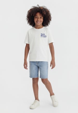 Bambino sorridente con capelli ricci che indossa una maglietta bianca "Youth Division", pantaloncini di jeans e scarpe da ginnastica bianche, in piedi su uno sfondo bianco semplice.