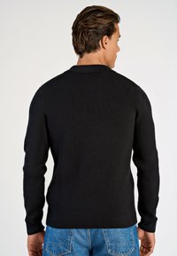 Schwarzer, gerippter Strickpullover mit langen Ärmeln und einem Kragen, der eine taillierte Passform und einen strukturierten Stoff aufweist, kombiniert mit blauen Jeans.
