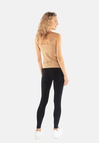 Dopasowany top na ramiączkach w kolorze brązowym, zestawiony z czarnymi legginsami. Modelka odwrócona tyłem, ukazująca gładką teksturę materiału oraz przylegający krój.