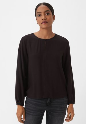 Black long-sleeve blouse with a round neckline, centered front pleat, and elasticized cuffs, paired with dark jeans. Smooth fabric texture.