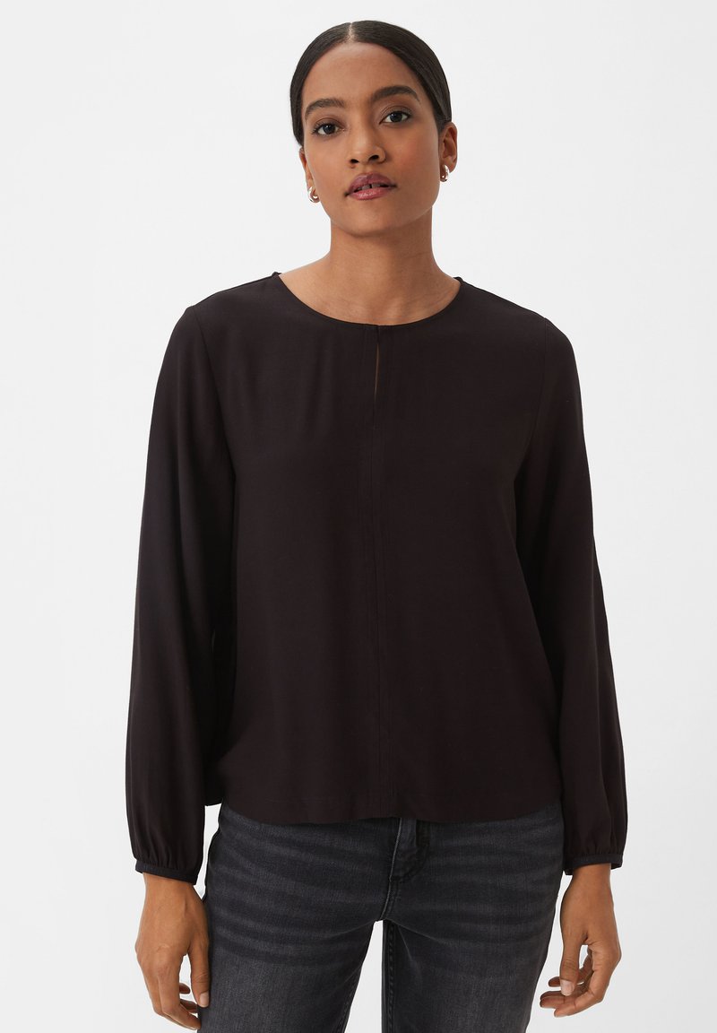 Black long-sleeve blouse with a round neckline, centered front pleat, and elasticized cuffs, paired with dark jeans. Smooth fabric texture.