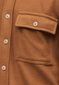 Καφέ πουκάμισο από μείγμα μαλλιού με θήκη στο στήθος, κλείσιμο με κουμπιά και μεγάλα στρογγυλά κουμπιά. Μαλακή υφή με ορατές λεπτομέρειες ραφών.