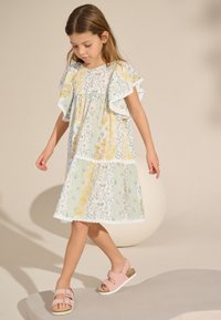 Kleid mit Blumenmuster in Gelb- und Grüntönen mit Rüschenärmeln, Spitzenbesatz und gestuftem Design. Rosa Sandalen mit Schnallenverzierung.