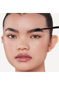 Close-up van het aanbrengen van make-up, met donkere wenkbrauwen die worden gekamd met een spoolie. Lichte huid, glanzende lippen en meerdere kleine oorbellen.