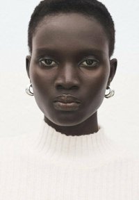 Primer plano de una persona de piel oscura, cabello corto, que lleva pendientes de aro plateados y un suéter de cuello alto blanco con canalé, mirando directamente al frente.