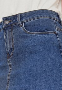 Närbild på midjan av blå denimjeans med framficka, bälteöglor och metallknapp, bärs med en instoppad vit topp.