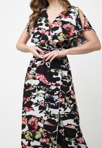 Femme portant une robe portefeuille fleurie noire et blanche avec des manches courtes à volants et des détails d'épaules ouvertes, posant avec les mains jointes à la taille.