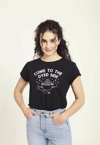 Camiseta negra de manga corta con el texto gráfico blanco "VEN AL LADO TEÑIDO" y un diseño que presenta un tazón y una cuchara. Combinada con jeans de color azul claro.