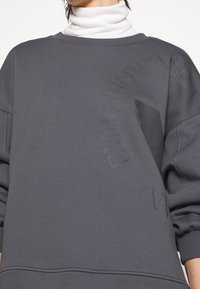 Sudadera gris con un corte holgado, que presenta detalles de logotipos en relieve y un acabado sutilmente texturizado. Capa de cuello alto visible.