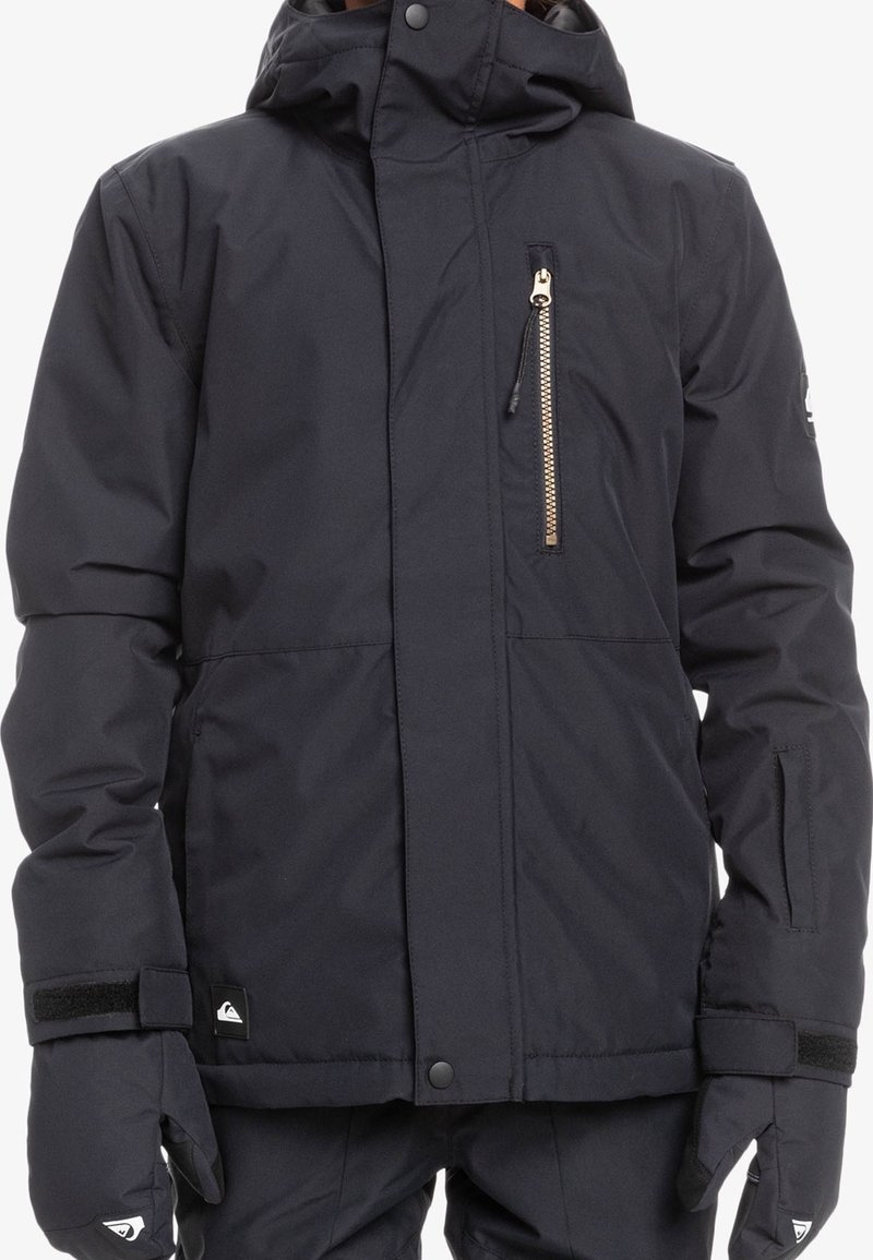 Personne portant une veste d'hiver isolante noire avec une poche poitrine zippée, une capuche réglable et des gants assortis sur fond blanc.